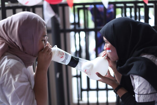 The image captures a balloon competition at Bahtera Adi Jaya Tekno during the celebration of Indonesian Independence Day. Two women are participating in the game, each blowing into a plastic bottle attached to a balloon, trying to inflate it as part of the competition. The festive atmosphere is highlighted by the decorations and the Indonesian flag face paint on one of the participants. This scene reflects the lively and engaging activities organized for the Independence Day celebration.