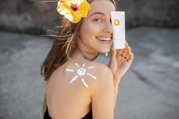Woman smiling holding a sunscreen bottle with a sun drawing made from sunscreen on her shoulder, symbolizing skin protection and care