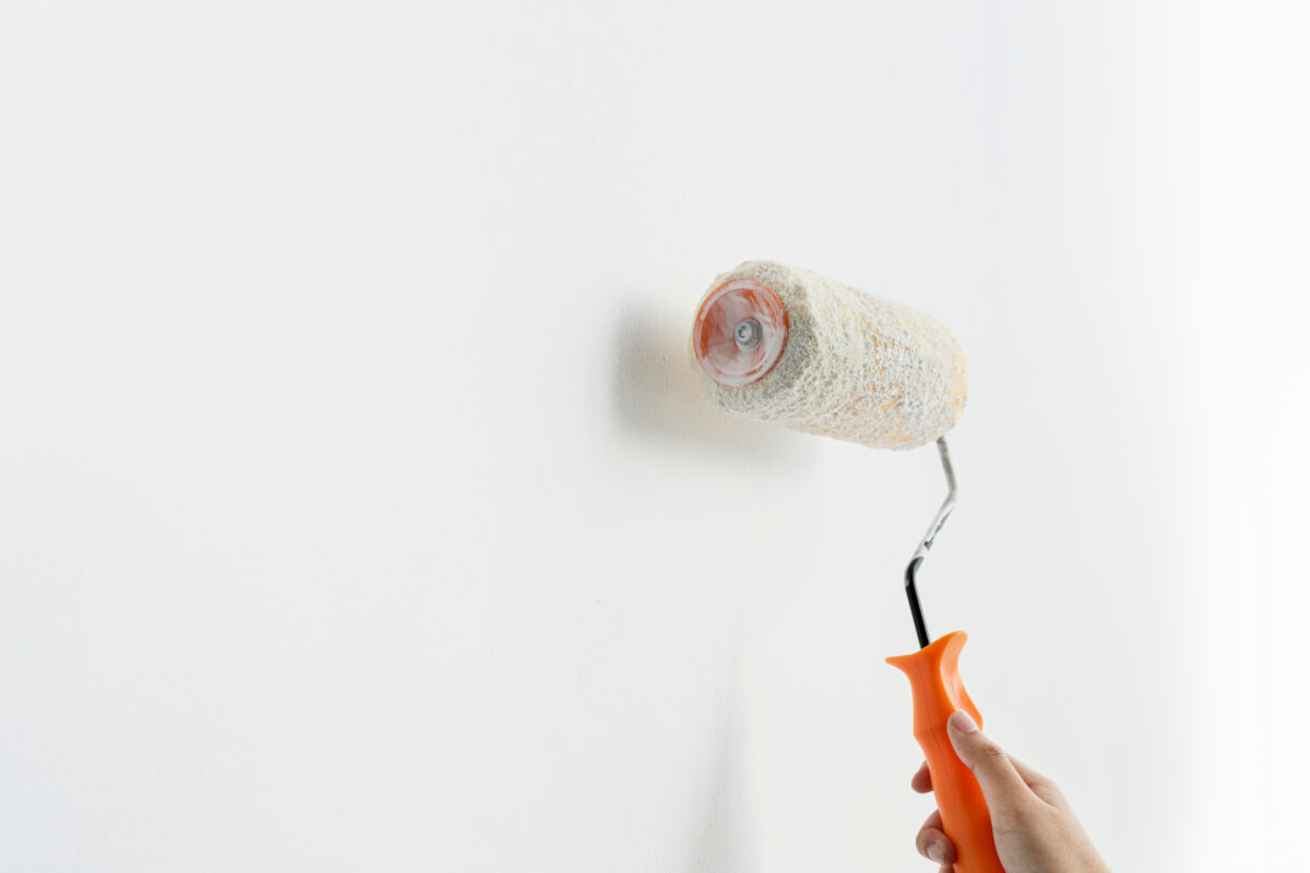 A hand holding an orange-handled paint roller applies white paint to a smooth wall. The roller is partially covered in paint, indicating that the painting process is in progress. Rheology modifiers in the paint enhance its application quality, contributing to a smooth and even finish.