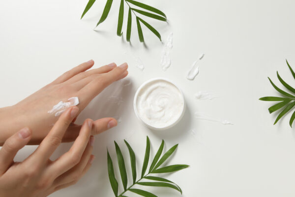 A pair of hands applying sustainable beauty cream from a jar, surrounded by green leaves, against a white background.