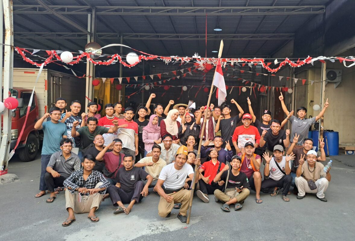 The image captures a celebration of Indonesia's Independence Day held at Bahtera Adi Jaya's Jatake location. The participants are gathered in front of the facility, decorated with red and white banners, symbolizing the national colors. The group is posing enthusiastically, reflecting the festive spirit of the occasion.