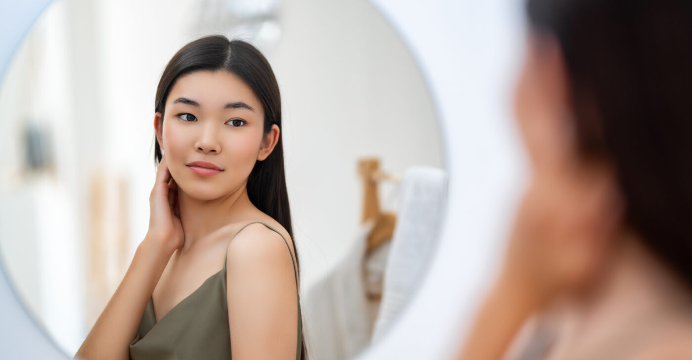 A young woman with smooth, clear skin, potentially representing relief from Atopic Dermatitis, gently touching her neck while looking at herself in a round mirror. She has long black hair and is wearing a green sleeveless top, standing in a softly lit, minimalistic room.