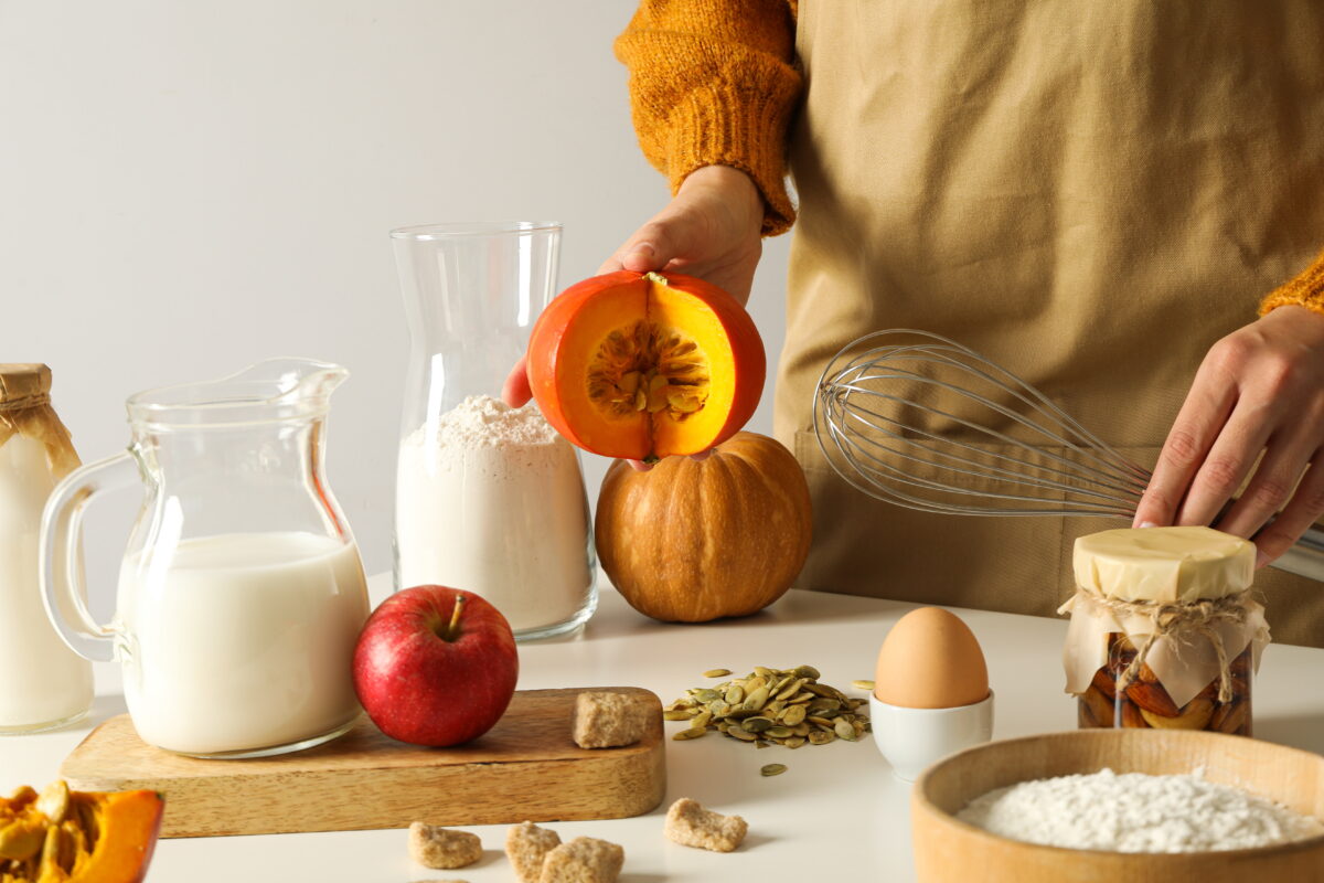 A person preparing ingredients for cooking, holding half of a pumpkin. Various ingredients like flour, milk, an apple, an egg, sugar cubes, seeds, and a jar of almonds are arranged on a table, with a whisk resting nearby, illustrating the concept of food fortification to enhance the nutritional value of meals.